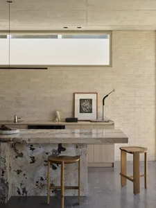 Modern minimalist kitchen with a large Calacatta marble island, two solid oak organic-shaped stools, light brick wall, and a framed abstract artwork under a high concrete ceiling.
