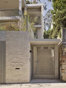 Modern minimalist house entrance with light-colored brick wall, vertical slatted metal gate, concrete structural elements, and lush greenery on upper terraces.