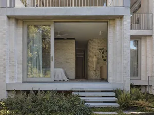 View from exterior into a modern bedroom featuring a curved light-colored brick wall, concrete structure, and large sliding glass doors opening to a garden.