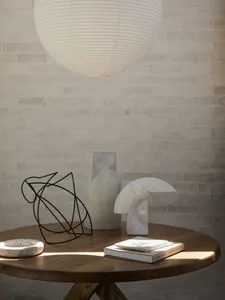 Still life arrangement on a round oak table featuring a black wire sculpture, travertine vase, marble lamp, speckled ceramic dish, stacked books, and a large white paper lantern hanging above against a white brick wall.