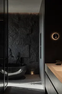 Modern dark minimalist bathroom featuring a glossy black freestanding tub against a textured natural stone wall, paired with a light wood floating vanity and dramatic wall sconce lighting.