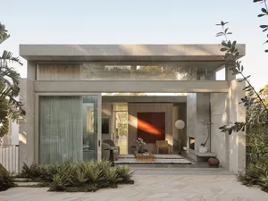 Modern concrete house with large glass sliding doors opening to a landscaped courtyard featuring ferns, wooden furniture, and a striking red abstract painting on the back wall.