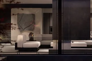 Modern minimalist living room featuring a large travertine feature wall, sculptural artwork, and neutral-toned modular sofas viewed through glass doors.