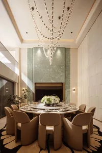 Luxurious modern dining room with a large round marble table, ten beige upholstered armchairs, a massive cascading crystal chandelier, sage green marble feature wall with fireplace, and patterned black and beige rug.
