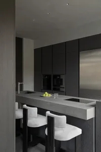 Modern minimalist kitchen with dark grey concrete island, white upholstered bar stools with dark wood frames, built-in black cabinetry, and stainless steel refrigerator.