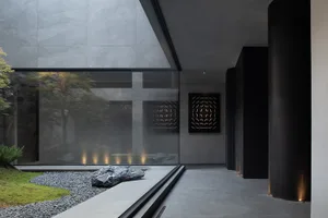 Modern minimalist interior entryway featuring large glass walls overlooking a Japanese zen rock garden with large basalt rocks, gravel, moss, and uplighting, finished with dark concrete walls and columns.