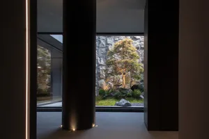 View from a dark minimalist interior hallway through a large opening to a Japanese-style courtyard featuring a stone wall, pine tree, and lush greenery.