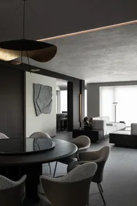 Modern minimalist dining area featuring a large round dark wood table, six textured boucle and leather chairs, an oversized black and gold sculptural pendant light, concrete walls, and a view into a light-colored living room with white sofas.