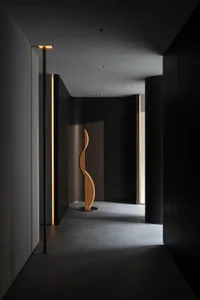 A minimalist dark hallway featuring a tall curved wooden sculpture, dramatic linear lighting, and matte black walls with concrete flooring.