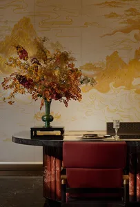 Round black marble dining table with red velvet upholstered chair, large autumnal floral arrangement in a green vase, and dramatic golden Chinese mountain-and-cloud mural on the wall.