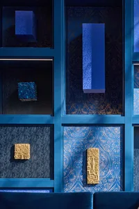 Close-up of a deep blue built-in shelving unit featuring patterned blue and gold wallpaper, blue and gold textured sculptural objects, integrated LED lighting, and a dark blue velvet sofa at the base.