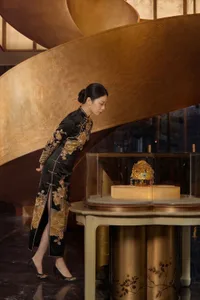 Elegant woman in traditional Chinese qipao viewing a golden opera headdress inside a glass display case set against a large curved bronze relief wall with mountain and cloud motifs.