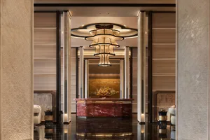 Elegant hotel lobby featuring a striking red marble reception desk, a large multi-tiered chandelier, glossy dark floor, and beige patterned wall panels.