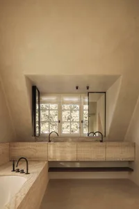 Modern minimalist bathroom featuring a long floating travertine vanity, black matte faucets, large mirror, and sloped ceiling with natural daylight from windows overlooking trees.