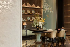 Luxurious contemporary Chinese dining room featuring a round fluted pedestal table, velvet dining chairs, a built-in mountain-shaped display shelf with porcelain, and a large floral mural with birds on a sage green wall.