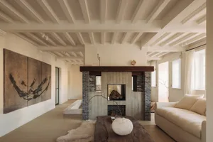 Modern minimalist living room featuring a central concrete and brick fireplace, whitewashed wooden ceiling beams, large abstract ink painting, and a rustic wooden coffee table on a sheepskin rug.