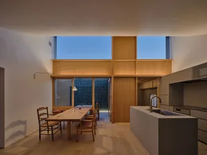 Modern open-plan dining area and kitchen featuring light oak cabinetry, a concrete kitchen island with integrated sink, wooden dining table with classic wooden chairs, and large windows overlooking a garden at dusk.