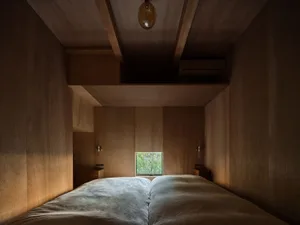 View from the foot of a bed looking into a compact plywood-clad bedroom with warm wooden walls, ceiling beams, wall sconces, and a small window showing greenery outside.