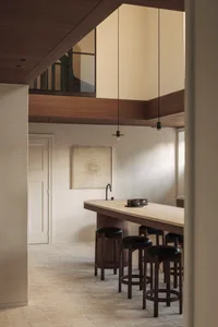 Modern minimalist kitchen with a long travertine stone island, dark wood cabinetry, black leather stools, and warm wood ceiling beams in a double-height space.