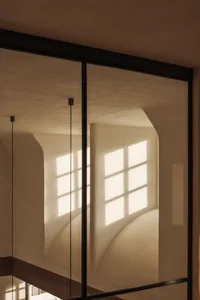 View through black steel-framed glass doors into a sunlit interior featuring smooth curved plaster walls and arched window openings.