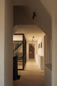 Long narrow hallway with light oak flooring, white walls, black steel and glass staircase on the left, built-in white storage cabinet on the right, and black cylindrical ceiling spotlights.