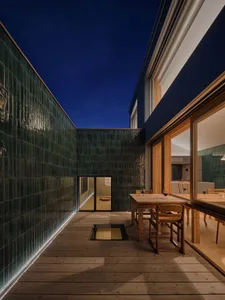 Night view of a modern courtyard with dark green glazed tile walls, wooden deck flooring, a built-in glass floor panel, and a wooden dining table set for four.