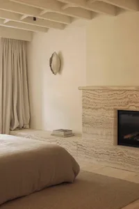 Modern minimalist bedroom featuring a large travertine fireplace and matching built-in ledge, neutral linen bedding, wooden floor, and exposed ceiling beams.