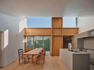 Modern open-plan dining and kitchen area featuring a curved concrete island, warm oak cabinetry, wooden dining table with rush-seat chairs, and large green-tiled outdoor view.