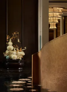 Luxury hotel entrance featuring a large ornate jade gourd sculpture on a dark wood base, set against dark wood paneling and polished black marble floor with mother-of-pearl inlay, with layered Chinese-style pendant lamps in the background.