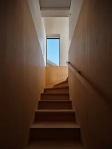 Looking up a narrow wooden staircase with light oak walls and handrail, natural daylight streaming from a tall vertical window at the top landing.