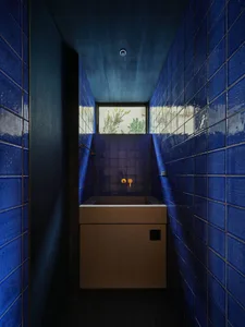 Narrow utility bathroom entirely clad in glossy cobalt blue tiles with a raw concrete sink and cabinet under a high window showing green foliage outside.