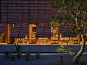 Night view of a modern Japanese-style house with a large built-in cedar wood bookshelf glowing with warm light, surrounded by a gravel garden and trees.
