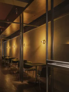 Narrow restaurant dining area with black tables and chairs lined against a textured wall, illuminated by warm brass wall sconces creating dramatic lighting and long shadows on a dark tiled floor.