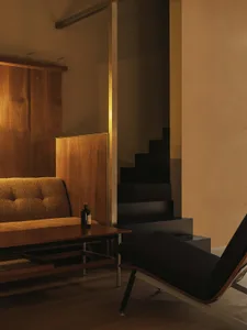 Modern living room corner featuring a mustard-yellow bouclé sofa, walnut wood paneling, a dark wooden coffee table, a black upholstered chaise lounge, and a black floating staircase in the background under warm lighting.