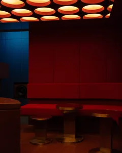Modern lounge area featuring curved red velvet banquette seating, metallic side tables, and a striking ceiling installation of glowing circular red and orange lights against deep blue and red walls.