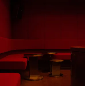 Modern lounge interior featuring a curved red velvet sofa, three cylindrical brushed metal nesting tables, and deep red padded walls under warm dramatic lighting.