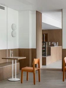 Modern minimalist cafe interior featuring light oak wood paneling on lower walls, white upper walls, a small square oak table with white base, and a bentwood oak chair.