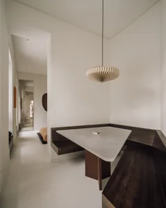 Modern minimalist corner dining area featuring a triangular concrete table, dark walnut L-shaped built-in bench, and a large beige paper pendant lamp in a bright white space.