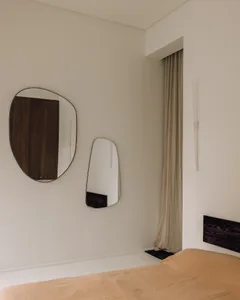 Minimalist bedroom corner featuring two asymmetrical mirrors with black walnut frames, beige linen curtains, and a walnut headboard against off-white walls.