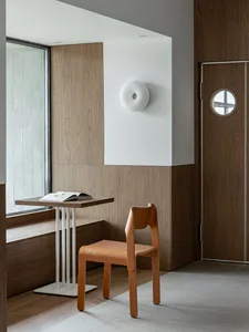 Modern minimalist reading nook featuring light oak chair, wall-mounted table, built-in window bench, and two-tone oak paneling with a circular wall sconce.