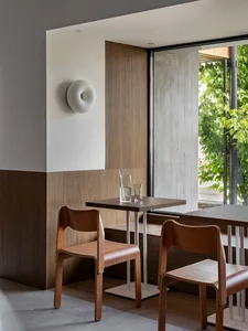 Modern minimalist cafe interior featuring walnut wood paneling, bentwood chairs, a small square table, and a large window overlooking green trees.