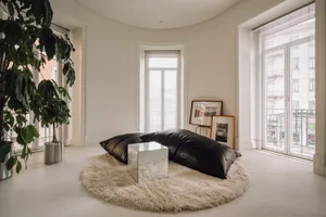 Bright minimalist living room with curved white walls, large windows with blinds, a circular cream sheepskin rug, black leather floor cushions, a mirrored acrylic side table, and a tall potted fiddle-leaf fig plant.