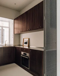 Modern minimalist kitchen featuring dark walnut wood cabinetry, stainless steel countertop, built-in oven, and a large window with white blinds.