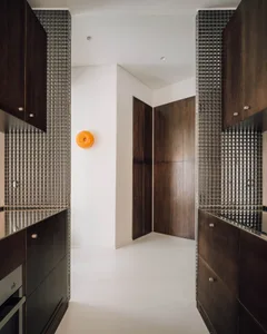 Modern narrow kitchen featuring dark wood cabinetry, silver mosaic tile walls, stainless steel countertops, and a bright orange circular wall sconce.