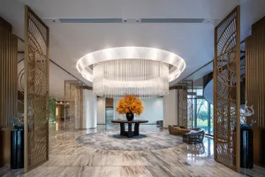 Spacious luxury hotel lobby with polished marble floors, a massive circular tiered chandelier, golden decorative room dividers, and a central round table with orange floral arrangement.