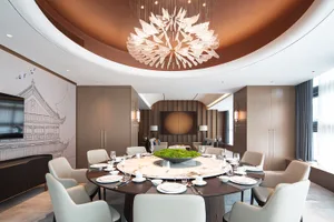 Luxurious circular dining room featuring a large white marble round table, eight cream upholstered chairs, dramatic crystal petal chandelier, warm wood paneling, and Chinese ink painting mural of a traditional pavilion.