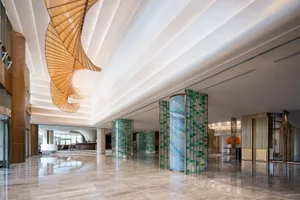 Spacious luxury hotel lobby with polished marble floors, blue and green mosaic pillars, and a large golden spiral chandelier hanging from a coffered ceiling.
