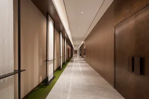 Long modern luxury hotel hallway with light gray marble floor, dark wood panel walls, beige textured wall covering, recessed ceiling lighting, and a strip of artificial grass along the left side.