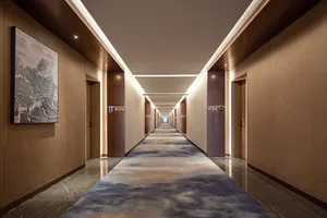 Long modern luxury hotel corridor with warm wood panel walls, dark marble flooring, patterned blue and beige carpet runner, recessed ceiling lighting, and large abstract mountain artwork on the left wall.