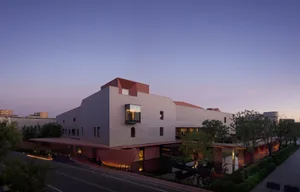 Exterior view of a contemporary concrete cultural center building at twilight, featuring bold geometric forms, cantilevered volumes, and warm interior lighting reflecting on a water feature.